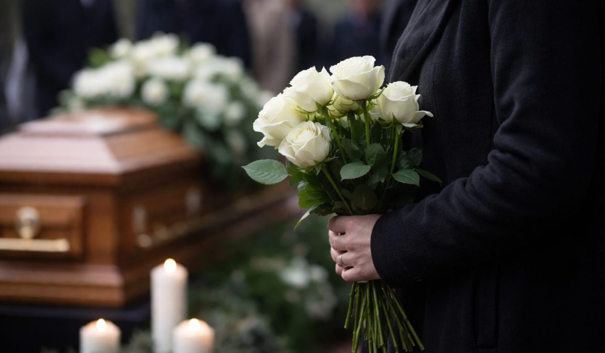 Personne en manteau noir tenant un bouquet de roses blanches devant un cercueil en bois, avec des proches en arrière-plan flou et des bougies allumées.